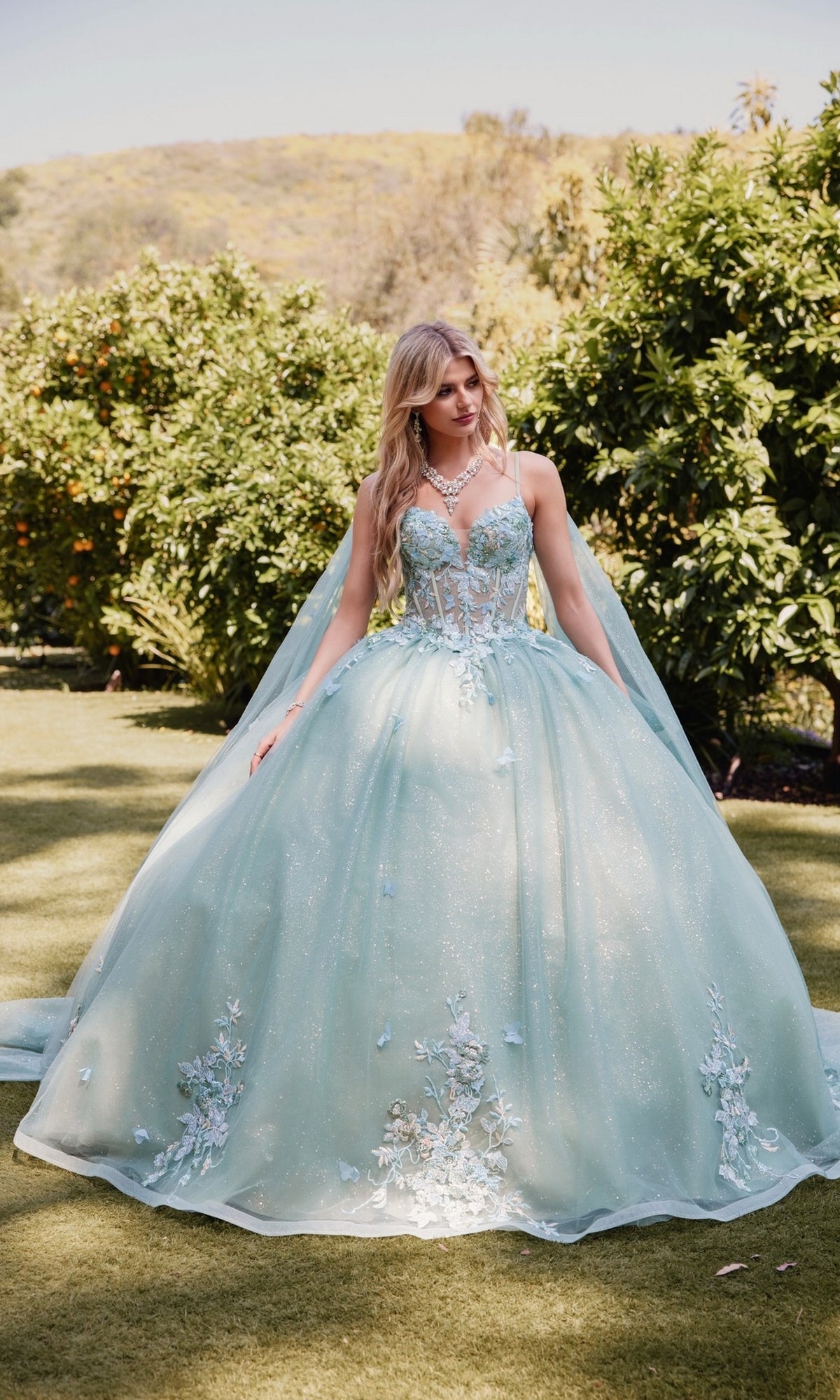 A woman in the Sage Green Quinceanera Dress: Juliet JT1466H, with a sheer corset bodice and floral embellishments, stands on grass in a garden surrounded by trees and rolling hills.